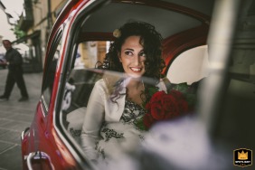 A bride sits in the backseat of a classic red Fiat 500 automobile, photographed at the bride's home before the wedding ceremony.